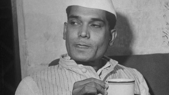 Photograph of a younger Jaya Prakash Narayan in a Gandhi cap and knitted waistcoat, seated, holding a cup