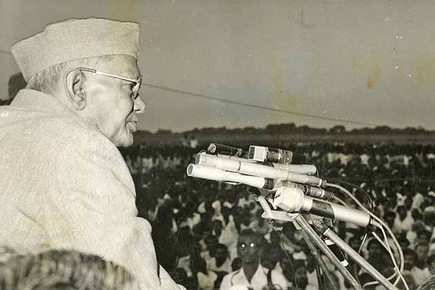 Photograph of Jaya Prakash Narayan in profile wearing a Gandhi cap, speaking into a bank of microphones before a vast seated crowd
