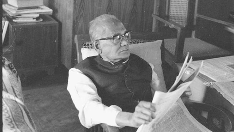 Photograph of Jaya Prakash Narayan seated in a chair in a room, holding a newspaper, with books stacked beside him