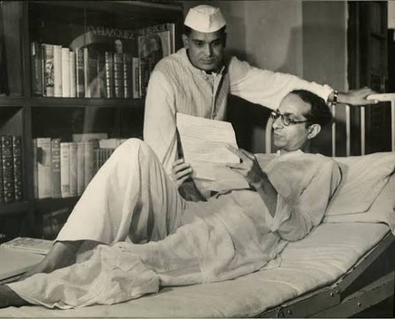 Photograph of Jaya Prakash Narayan reclining on a bed reading a sheaf of papers, with a man standing beside him and bookshelves behind