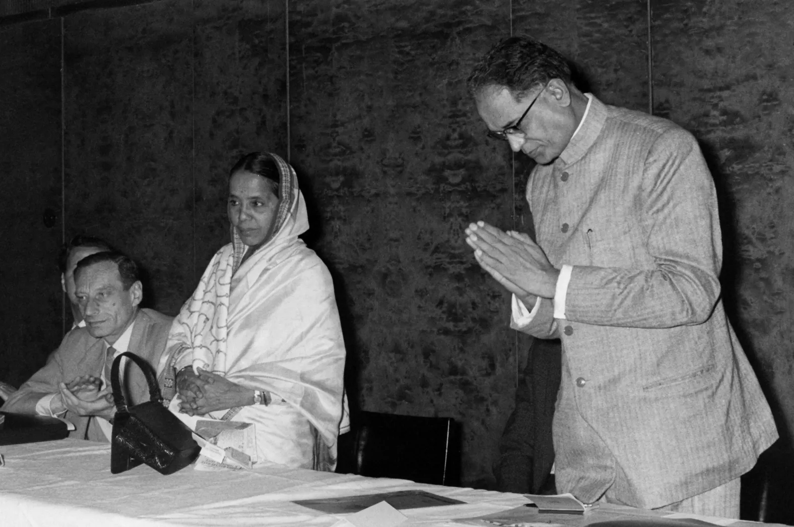 Photograph of a man in a kurta greeting with folded hands beside a seated woman in a sari at a conference table
